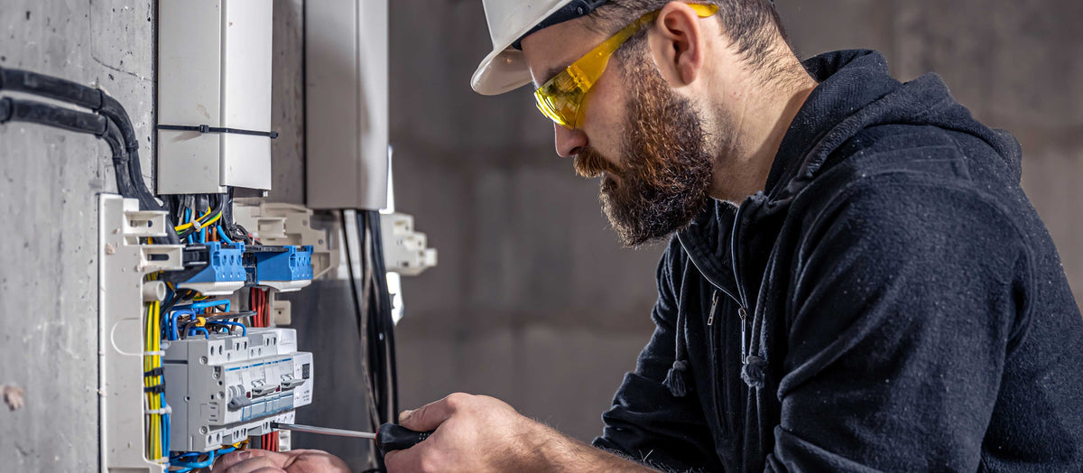 Elektriker mit Schutzhelm und Schutzbrille verdrahtet Reiheneinbaugeräte in einem geöffneten Verteilerschrank
